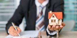 Homme en costard qui tient la maquette d'une maison et des clefs dans sa main, avc quelques pièces de monnaie posées sur le bureau