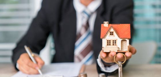 Homme en costard qui tient la maquette d'une maison et des clefs dans sa main, avc quelques pièces de monnaie posées sur le bureau