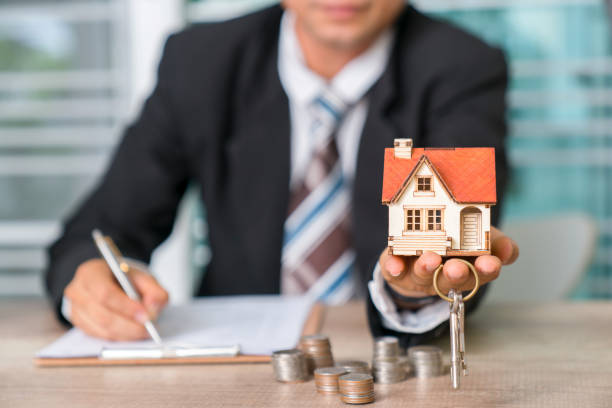 Homme en costard qui tient la maquette d'une maison et des clefs dans sa main, avc quelques pièces de monnaie posées sur le bureau